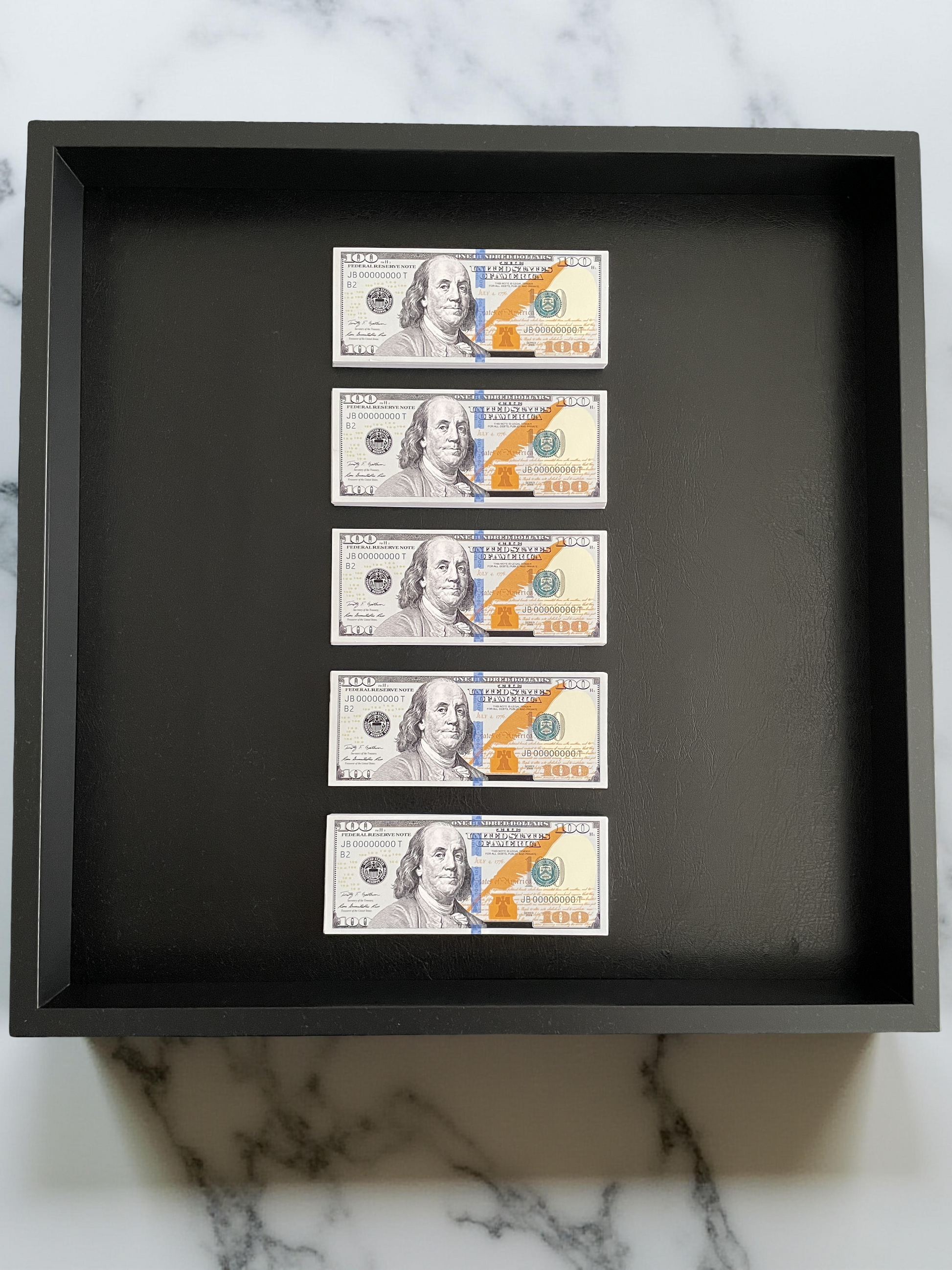 Five U.S. hundred-dollar bills arranged vertically in a black shadow box on a marble surface.