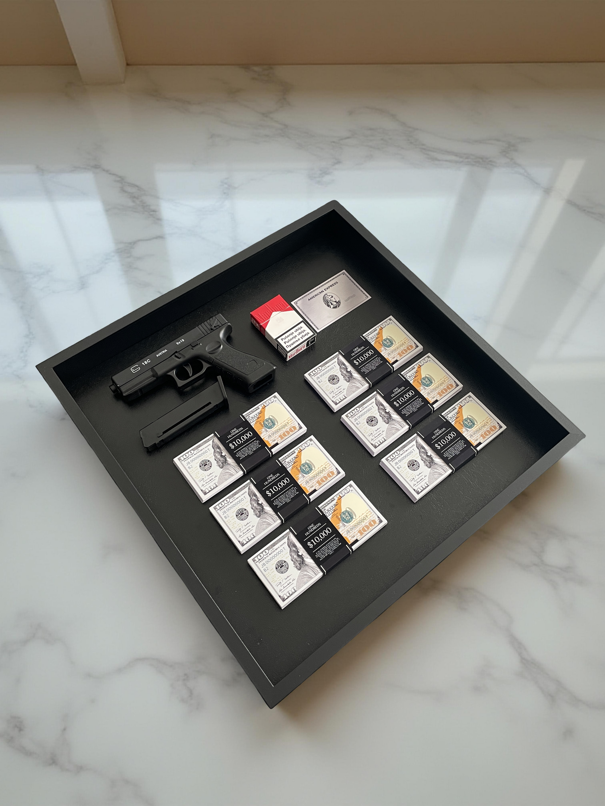 A black shadowbox showcasing a Glock, Marlboro cigarettes, an Amex card, and six stacks of USD.