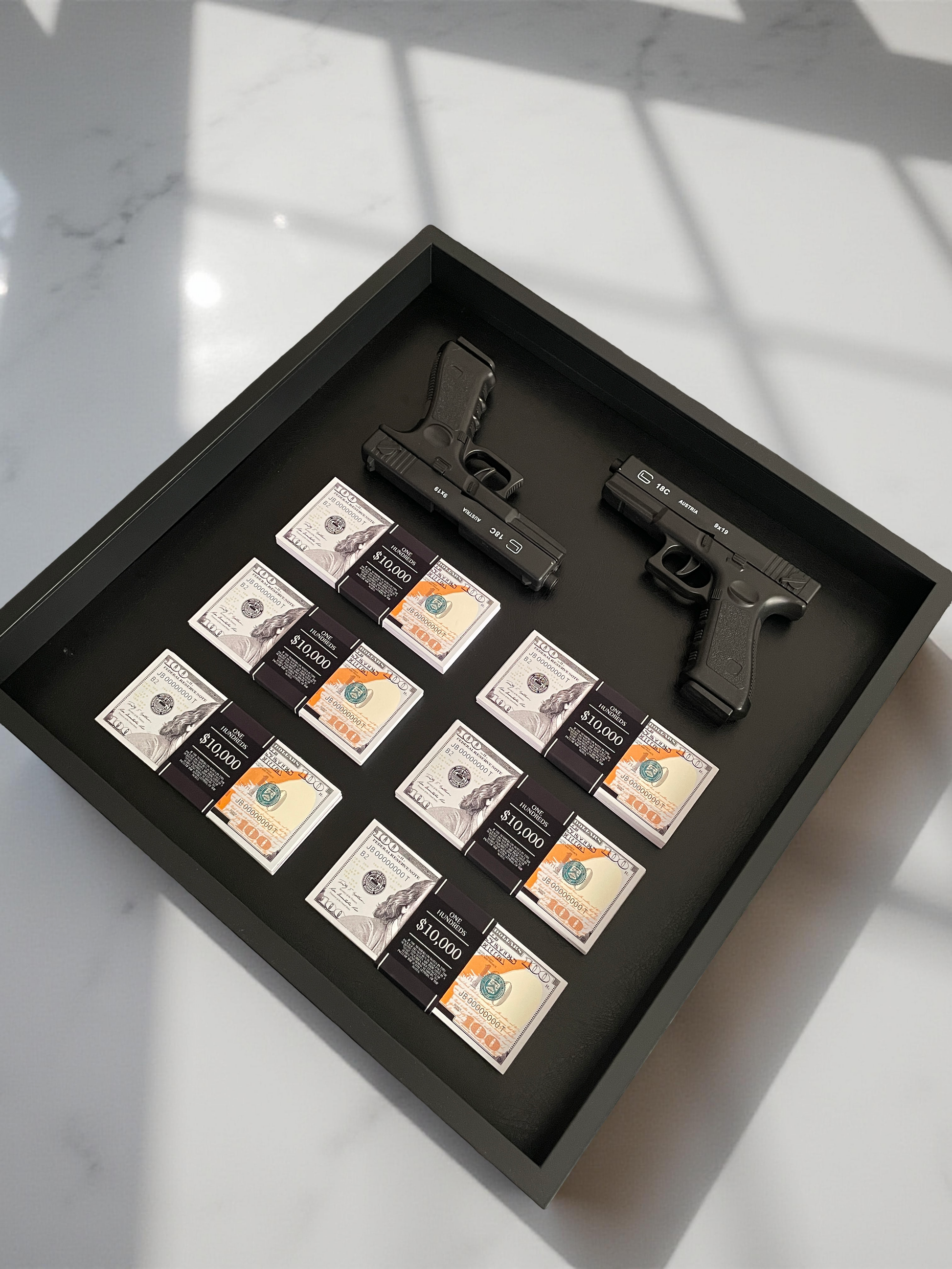 Two Glocks displayed alongside stacks of US dollar bills, both encased in sleek black shadowboxes.